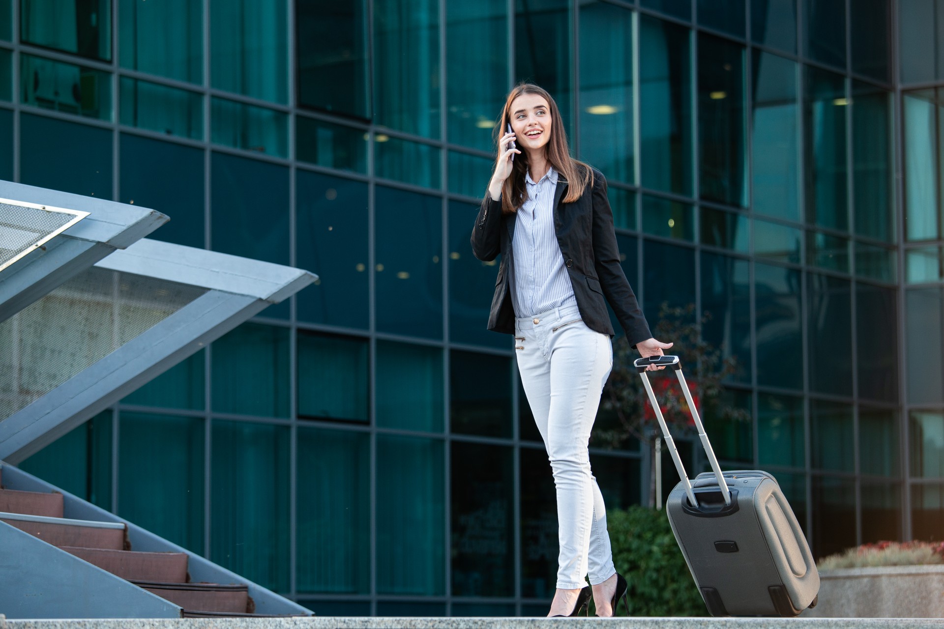 Pretty smiling female flight attendant carrying baggage going to airplane in the airport. Business woman at international airport looking at her mobile phone